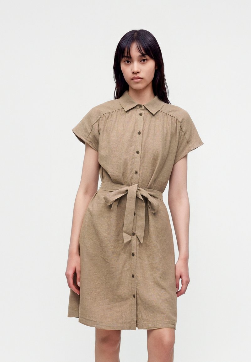 Jeune femme portant une robe beige à manches courtes boutonnée, avec un col et une ceinture en tissu nouée, debout devant un fond clair uni.