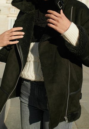 Dark green shearling jacket with a zippered front, black fleece lining, and a contrasting white ribbed sweater underneath.