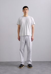 Hellgraues Baumwoll-T-Shirt, lockere Passform, und hellgraue Hose mit geradem Schnitt. Schwarze Schuhe mit Holzsohle. Einfarbiger Hintergrund.