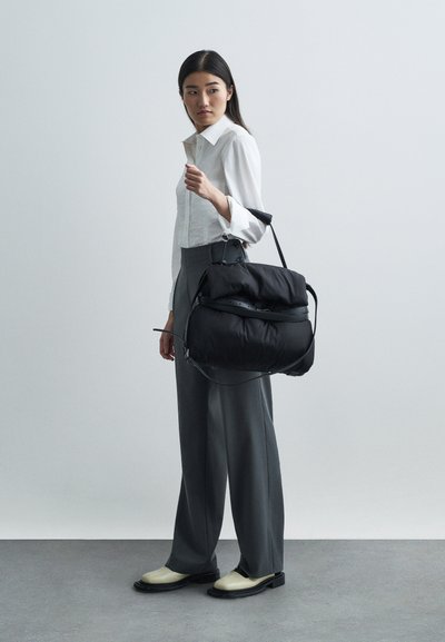 Sac à puffer noir avec une surface texturée, une courte poignée et une bandoulière. Le modèle porte une chemise blanche et un pantalon large gris avec des chaussures noires.