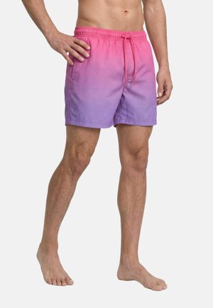 Hombre con bañador tipo shorts con degradado de rosa a púrpura, cintura elástica y cordón, de pie descalzo con una mano en la cadera.