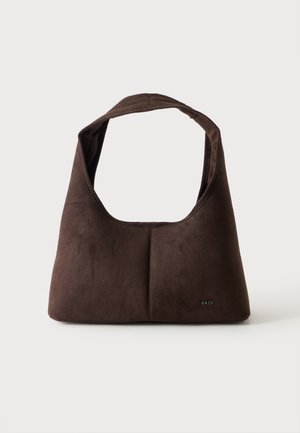 Sac bandoulière en cuir suédé marron foncé avec une large anse et un marquage subtil dans le coin inférieur droit sur un fond uni.