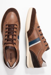 Baskets en cuir marron et daim avec une bande blanche et bleue, bout arrondi et fermetures à lacets. Elles présentent une semelle en caoutchouc texturée.