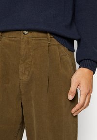 Homme portant un pantalon en velours côtelé marron et un pull bleu marine, la main droite détendue le long du corps, sur un fond uni.