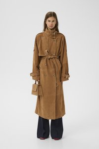 Cappotto trench in suede marrone con colletto alto, cintura e bottoni. Abbinato a pantaloni a gamba larga a righe e una piccola borsa strutturata.