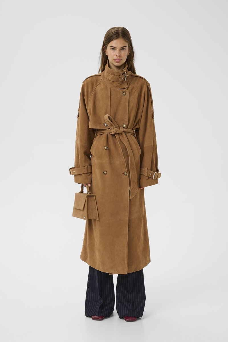 Cappotto trench in suede marrone con colletto alto, cintura e bottoni. Abbinato a pantaloni a gamba larga a righe e una piccola borsa strutturata.