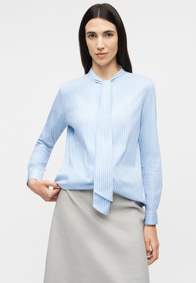 Γυναίκα που φοράει μπλούζα με μακριά μανίκια, ανοιχτού γαλάζιου χρώματος, με κάθετες ρίγες και λεπτομέρεια δεσίματος στο λαιμό, καθώς και φούστα ανοιχτού γκρι χρώματος.