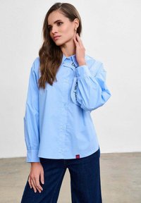 Femeie cu păr lung și castaniu, purtând o cămașă supradimensionată albastru deschis cu detaliu de guler în dungi și blugi în culoare albastru închis, stând în picioare pe un fundal simplu.
