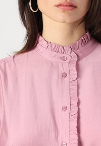 Lys rosa bluse med volangkrage, frontlukking med knapper og glatt tekstur. Knapper som er litt blanke og matcher stoffets farge.