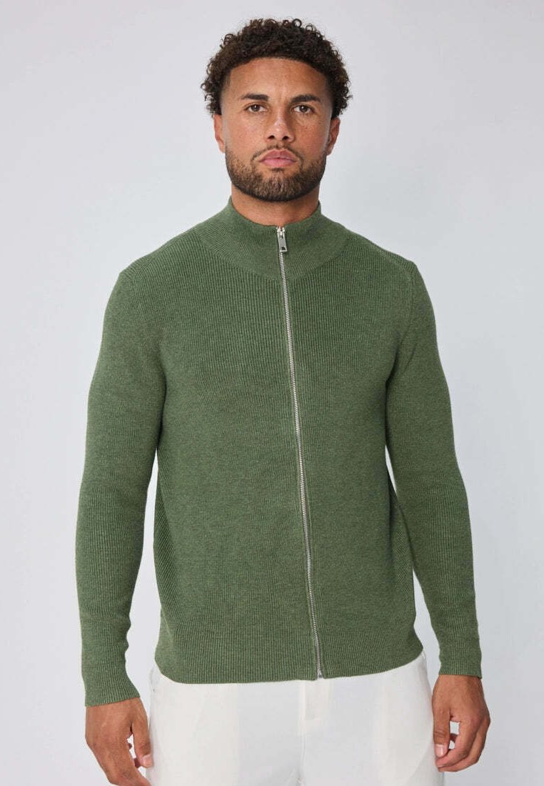 Homme aux cheveux bouclés et à la barbe portant un pull zippé à côtes vert et un pantalon blanc, faisant face avec une expression neutre.