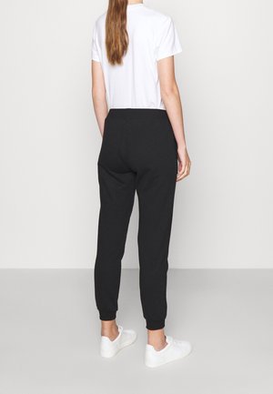 Persoon staat met de rug naar voren, draagt een wit T-shirt met korte mouwen, zwarte joggingbroek en witte sneakers op een effen achtergrond.