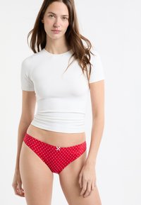 Roter Bikini mit weißen Tupfen und einer weißen Schleifen-Akzentuierung, gefertigt aus glattem, weichem Stoff mit niedrig sitzendem Design, kombiniert mit einem enganliegenden weißen Top.