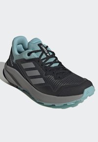 Chaussure de sport noire et turquoise avec tige en mesh, accents gris et semelle en caoutchouc. Elle dispose de lacets et de trois bandes latérales. Marque Terrex visible.