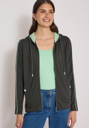 Kvinde iført en sort hættetrøje med lynlås og mintgrøn foer over en mintgrøn tanktop og blå jeans, smilende med afslappede hænder.