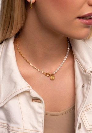 Goldkette mit einer Perlenstrang und einem runden Anhänger mit strukturierten Details. Getragen über einem hellen Oberteil unter einer Jacke.