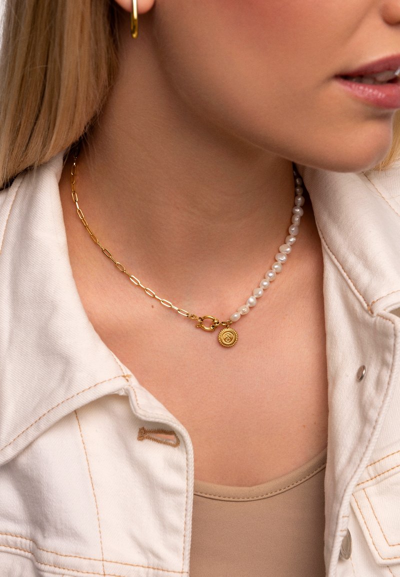 Collar de cadena de oro con una hebra de perlas y un colgante redondo con un acabado texturizado. Lleva puesto sobre una blusa de color claro debajo de una chaqueta.