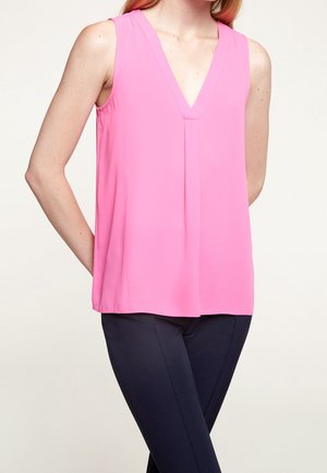 Bez rękawów, jasnoróżowa bluzka w kształcie litery V zestawiona z dopasowanymi czarnymi spodniami, noszona przez osobę z jedną ręką za plecami.