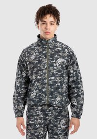 Camouflagejakke og bukser i nuancer af grå og sort. Har en høj krave, lynlås lukning, elastiske manchetter og hvide accenter.