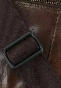 Brown leather bag with a dark strap and a black rectangular buckle; features visible stitching and a textured surface.