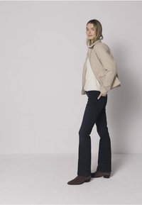 Beige jas met een gestructureerd ontwerp, daaronder een witte trui, donkere uitlopende jeans en bruine enkellaarzen, tegen een neutrale achtergrond.