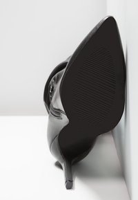 Zapato de tacón alto negro con punta afilada, parte superior de cuero liso y suela de goma texturizada, apoyado sobre una superficie blanca.