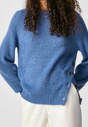 Kobieta ubrana w niebieski dzianinowy sweter z guzikami po bokach i białe spodnie, stoi z rękami za plecami.