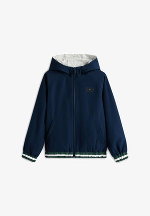 Lächelnder Junge mit marineblauer Mütze, marineblauer Zip-up-Jacke, weißem Polohemd mit Streifen, weißen Shorts mit grünem Logo, marineblauen Socken und marineblauen Turnschuhen.