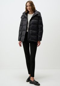 Doudoune noire avec fermeture éclair, dotée d'un col haut, d'un motif matelassé horizontal et de poches pression sur le devant. Portée avec un pantalon sombre.