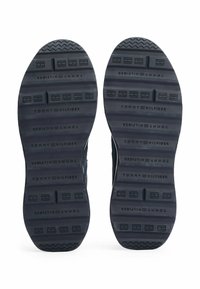 Suole di sneaker in gomma nera con un pattern di grip testurizzato e branding in rilievo che recita "TOMMY HILFIGER". Progettate per comfort e trazione.