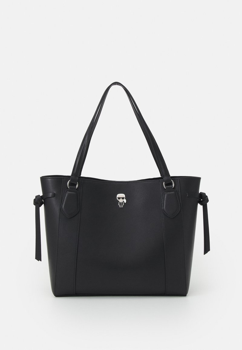 Sac fourre-tout en cuir noir avec deux bretelles d'épaule, des détails noués sur les côtés, et un petit emblème argenté en forme de caractère au centre avant.