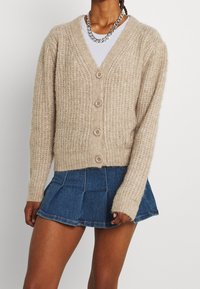 Beige knitted cardigan with a v-neck, five buttons down the front, and ribbed texture, paired with a denim mini skirt.