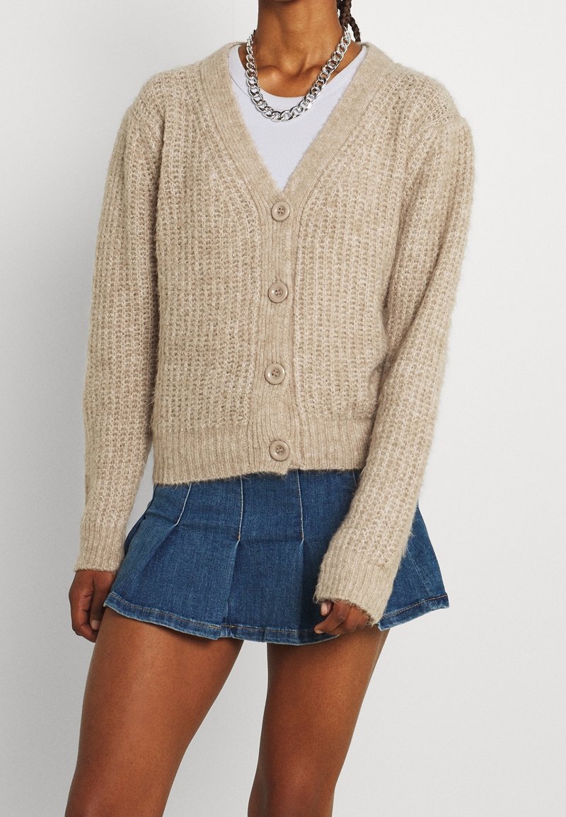 Beige knitted cardigan with a v-neck, five buttons down the front, and ribbed texture, paired with a denim mini skirt.