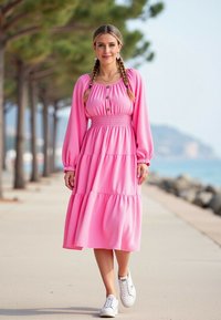 Jasnoróżowa, warstwowa maxi sukienka z długimi rękawami, marszczonym pasem i guzikami przy dekolcie, zestawiona z białymi trampkami na chodniku.