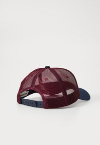 Casquette rouge en mesh à l'arrière avec une visière bleu marine, fermeture réglable par pression et petits trous de ventilation. Texture lisse sans motifs visibles.