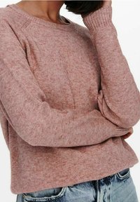 Pull en maille rose avec col rond, surface texturée, épaules tombantes et poignets côtelés. Associé à un jean en denim bleu déchiré.