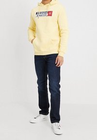 Sudadera de color amarillo claro con un logo en el frente, combinada con jeans de color azul oscuro y zapatillas blancas. La sudadera cuenta con cordones y un bolsillo delantero.