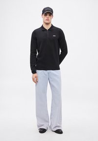 Črna polo majica z dolgimi rokavi in zadrgo na ovratniku, z logotipom na prsih, kombinirana s svetlo modrimi širokimi kavbojkami in črnimi čevlji, nošena s črno kapo.