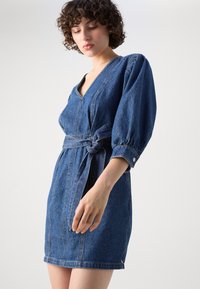 Denimkjole med v-utringning, korte pufferm og knyttet midje. Har kontrastsøm og hem over kneet.
