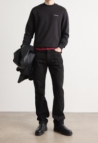 Felpa nera con logo bianco, indossata sopra una maglietta borgogna. Jeans neri e stivali di pelle neri completano l'outfit.