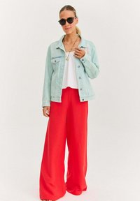 Jasnoniebieska dżinsowa kurtka, biała bluzka i jaskrawoczerwone szerokie spodnie. Modelka nosi czarne okulary przeciwsłoneczne i naszyjnik. Gładkie tekstury materiału.