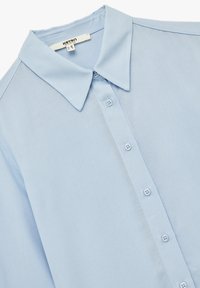Lyseblå skjorte med krave, der har en glat tekstur og seks matchende knapper foran. Casual design.