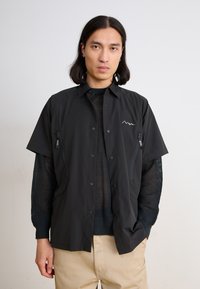 Black short-sleeved shirt featuring a zippered pocket, a mesh inner layer, and subtle mountain logo detail on the left chest.