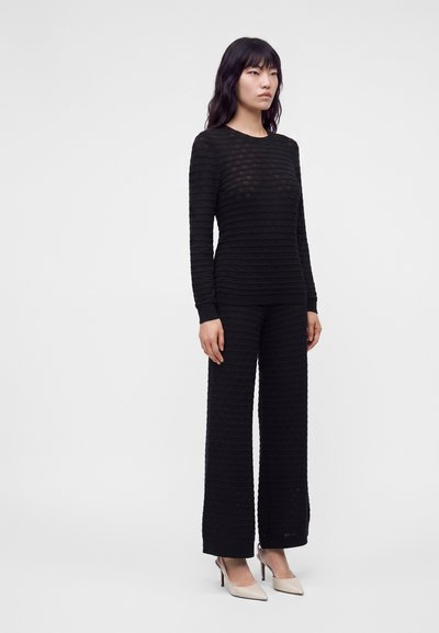 Femme debout portant un haut à manches longues texturé noir et un pantalon large avec des chaussures à talons beiges sur un fond blanc uni.