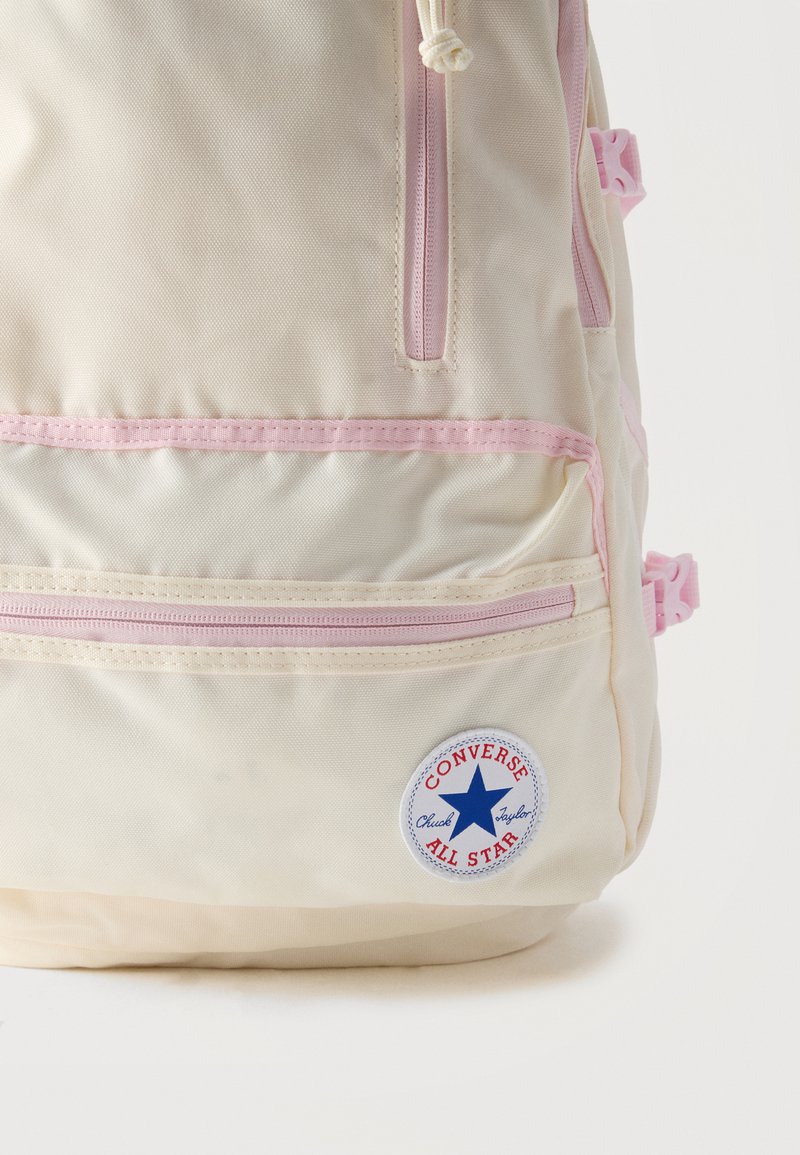 Zaino Converse color crema con cerniere rosa, due tasche frontali, fibbie laterali e una toppa rotonda con il logo Chuck Taylor All Star.