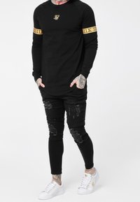 Schwarzes Langarmshirt mit goldenen Akzenten, kombiniert mit zerrissenen schwarzen Skinny-Jeans und weißen Sneakers mit goldenen Details.