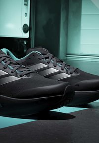 Paire de chaussures de sport noires et bleu sarcelle avec des bandes argentées placée sur un sol à carreaux près de murs bleu sarcelle et un appareil blanc.