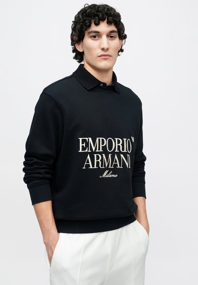 Czarna bluza z napisem "EMPORIO ARMANI Milano" w jasnozłotym kolorze. Okrągły dekolt z delikatnym kołnierzykiem i dopasowanymi mankietami. Gładka faktura.
