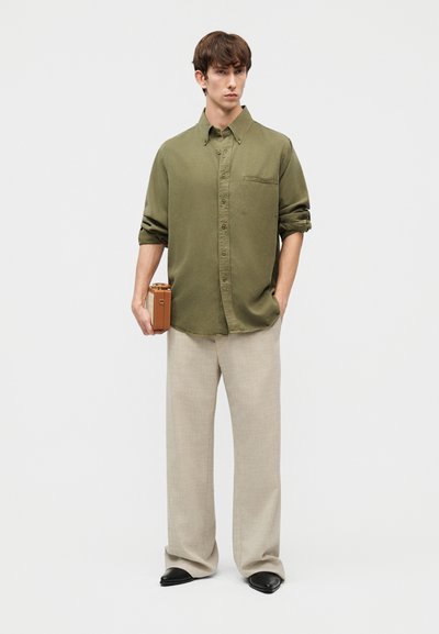 Άνδρας που φοράει πουκάμισο σε ελιά χρώμα με κουμπιά και μπεζ παντελόνι, κρατώντας μια μικρή καφέ δερμάτινη τσάντα μπροστά σε μια απλή λευκή επιφάνεια.
