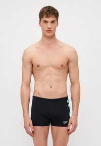 Černé plavky s těsným střihem, s vertikálními pruhy v námořnické modré a zelené barvě po stranách a logem Speedo na spodní přední části.
