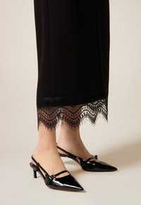 Falda larga negra con ribete de encaje que presenta patrones ondulados. Par de tacones slingback de charol negro con punta puntiaguda y tacón delgado.